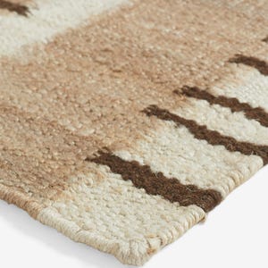 May include: A woven rug featuring a textured surface with alternating horizontal bands of cream, tan, and dark brown. The pattern consists of thick, solid-colored stripes interspersed with sections of darker brown vertical fringe-like accents.