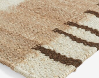 May include: A woven rug featuring a textured surface with alternating horizontal bands of cream, tan, and dark brown. The pattern consists of thick, solid-colored stripes interspersed with sections of darker brown vertical fringe-like accents.