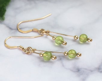May include: A pair of gold earrings with a simple design. Each earring has a gold hook, a small gold bead, a green gemstone, and another gold bead. The earrings are presented on a marble surface.
