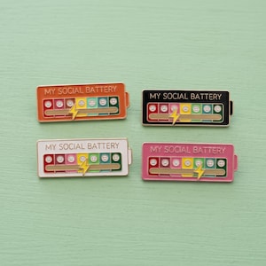 May include: Four rectangular enamel pins shaped like batteries, each featuring the text MY SOCIAL BATTERY and a row of faces ranging from sad to happy. The pins come in orange, black, white, and pink, with a yellow lightning bolt icon.
