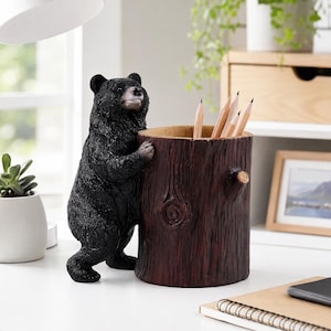 May include: A black bear figurine standing on its hind legs, leaning against a cylindrical, dark brown tree stump pencil holder. The stump features a textured bark pattern and a small knot, containing several wooden pencils inside the hollow top.