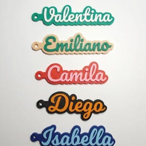 May include: Five custom name keychains with a layered design. The names Valentina, Emiliano, Camila, Diego, and Isabella are displayed in various color combinations including teal, white, beige, green, red, pink, black, orange, blue, and light blue.