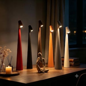 May include: Six tall, slender, conical table lamps in red, black, dark brown, orange, gray, and white. Each lamp features a small, angled light source at the top. The lamps are arranged in a row on a wooden surface alongside a small sculpture.