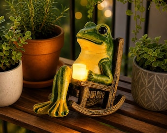 May include: A green and yellow frog figurine sitting in a small brown wooden rocking chair. The frog has large black eyes and is holding a small, glowing, cylindrical light that emits a warm yellow illumination from its centre.