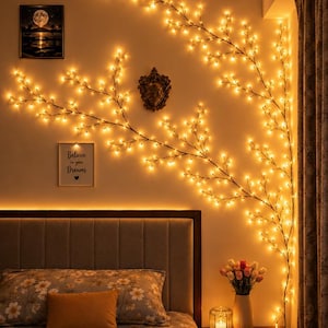 Op de afbeelding: Een decoratieve takvormige lichtsnoer met talloze kleine, warmgekleurde led-lampjes langs dunne, donkerbruine stelen. De flexibele lichtstreng is aan de muur bevestigd en vormt een uitgestrekt, gloeiend silhouet dat op een boom lijkt.
