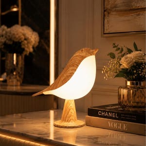 Puede incluir: Una lámpara de mesa con forma de pájaro que presenta un acabado de veta de madera clara en la cabeza y la base, con un cuerpo blanco liso y brillante que emite una luz suave y cálida. La lámpara se apoya en una base circular y tiene un diseño curvo.