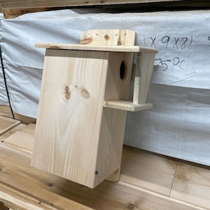 May include: A wooden birdhouse with a small opening for birds to enter. The birdhouse is made of natural wood and has a simple design. The birdhouse is 9 inches by 8 inches and is part of a set of 25.