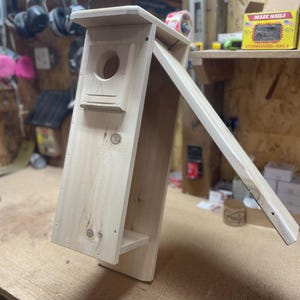 May include: A wooden birdhouse with a hinged front panel, revealing the interior. The birdhouse is made of light-colored wood and has a small round opening for birds to enter.