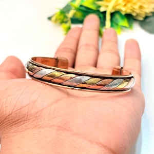 以下が含まれることがあります： ツイストロープデザインの3色カフブレスレット。銅、真鍮、銀色の3つの異なる金属バンドを編み込み、手首の周りに連続した質感のあるパターンを作り出しています。
