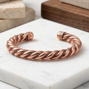 May include: A copper cuff bracelet featuring a twisted rope design with a polished metallic finish. The bracelet is an open-ended circular band with rounded, textured end caps, showcasing a warm reddish-brown color throughout the entire piece.