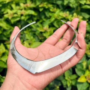 May include: A silver-toned metal choker necklace featuring a smooth, polished surface and a distinct V-shaped centre point. The rigid, open-ended collar design has a reflective finish that catches light, creating a sleek and uniform appearance.