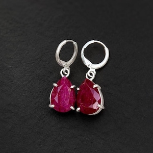 May include: A pair of silver hoop earrings featuring faceted, pear-shaped deep red gemstones. Each stone is secured in a four-prong setting that hangs from a circular silver hoop. The earrings have a polished metallic finish and a vibrant red hue.