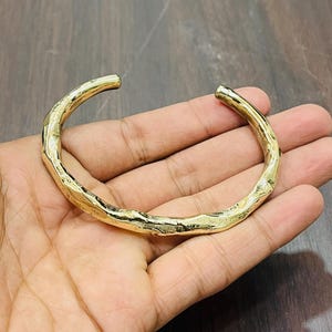 Op de afbeelding: Een goudkleurige metalen armband met een open ontwerp en een getextureerd, onregelmatig oppervlak dat lijkt op een natuurlijke tak of twijg. De armband heeft een gepolijste, reflecterende afwerking en een gebogen, cirkelvormige vorm.