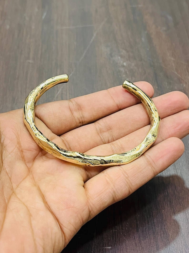 Op de afbeelding: Een goudkleurige metalen armband met een open ontwerp en een getextureerd, onregelmatig oppervlak dat lijkt op een natuurlijke tak of twijg. De armband heeft een gepolijste, reflecterende afwerking en een gebogen, cirkelvormige vorm.