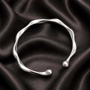 May include: A silver-toned open cuff bracelet featuring a continuous, undulating, wave-like design. The bracelet has a polished, reflective surface and terminates at both ends with a smooth, rounded, spherical bead detail.