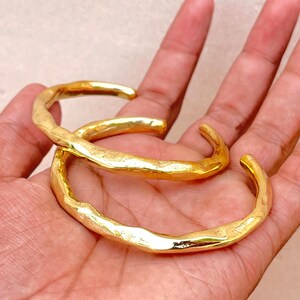 K&ouml;nnte beinhalten: Zwei goldfarbene Manschettenarmb&auml;nder aus Metall mit einer strukturierten, unebenen Oberfl&auml;che. Jedes Armband hat ein offenes Design mit einer polierten, reflektierenden Oberfl&auml;che, die das Licht einf&auml;ngt und ein welliges, organisches Aussehen erzeugt.