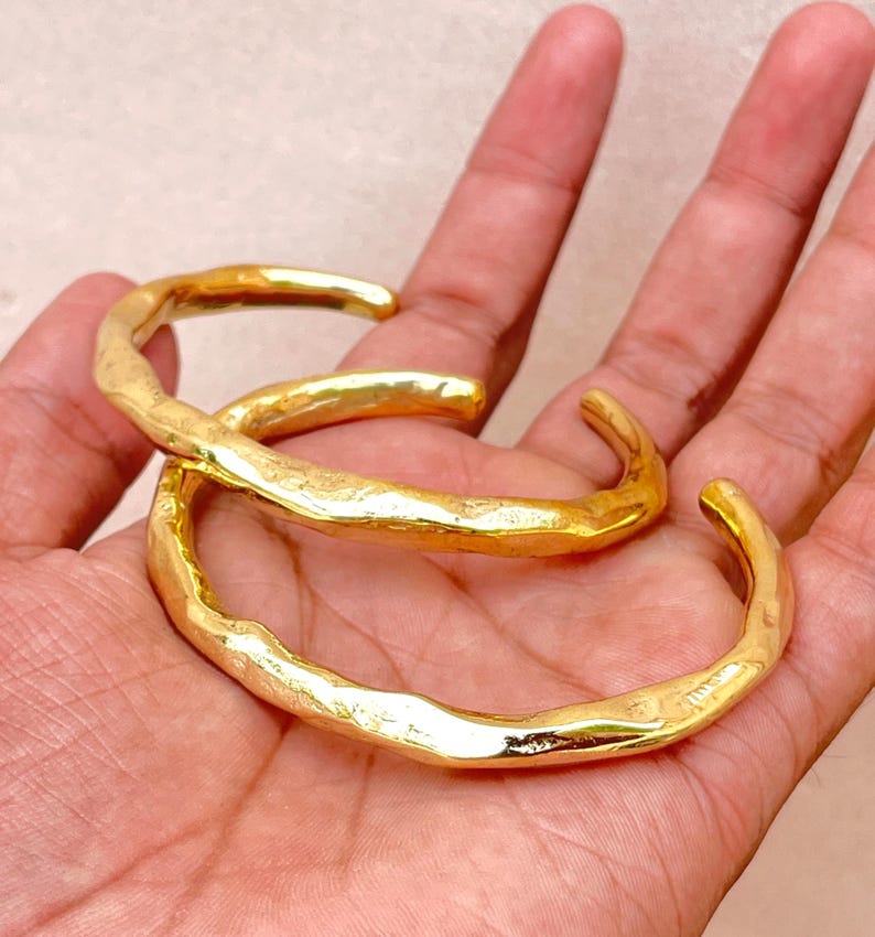 K&ouml;nnte beinhalten: Zwei goldfarbene Manschettenarmb&auml;nder aus Metall mit einer strukturierten, unebenen Oberfl&auml;che. Jedes Armband hat ein offenes Design mit einer polierten, reflektierenden Oberfl&auml;che, die das Licht einf&auml;ngt und ein welliges, organisches Aussehen erzeugt.