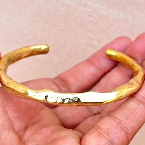 K&ouml;nnte beinhalten: Ein goldfarbenes Armband in Manschettenform mit polierter, reflektierender Oberfl&auml;che und einer unregelm&auml;&szlig;igen, organischen Textur. Das Armband hat ein offenes Design mit abgerundeten, glatten Enden und einem unebenen Metallband.