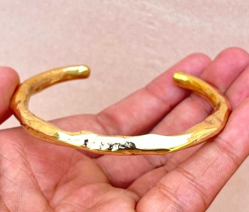 K&ouml;nnte beinhalten: Ein goldfarbenes Armband in Manschettenform mit polierter, reflektierender Oberfl&auml;che und einer unregelm&auml;&szlig;igen, organischen Textur. Das Armband hat ein offenes Design mit abgerundeten, glatten Enden und einem unebenen Metallband.