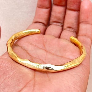 K&ouml;nnte beinhalten: Ein goldfarbenes Armband mit einer unregelm&auml;&szlig;igen, organischen und strukturierten Oberfl&auml;che. Das Metallband weist eine durchgehende, wellenf&ouml;rmige Form mit einer polierten Oberfl&auml;che auf, die das Licht auf den unebenen Kanten reflektiert.