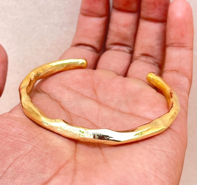K&ouml;nnte beinhalten: Ein goldfarbenes Armband mit einer unregelm&auml;&szlig;igen, organischen und strukturierten Oberfl&auml;che. Das Metallband weist eine durchgehende, wellenf&ouml;rmige Form mit einer polierten Oberfl&auml;che auf, die das Licht auf den unebenen Kanten reflektiert.