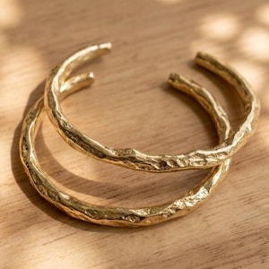 May include: Two gold-toned metal cuff bracelets featuring a textured, uneven surface that mimics the appearance of organic tree bark. Each bracelet is an open-ended circular band with a polished finish that reflects light across its irregular ridges.