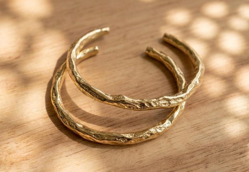 Op de afbeelding: Twee goudkleurige metalen armbanden met een getextureerd, onregelmatig oppervlak dat lijkt op de textuur van organische boomschors. Elke armband is een open cirkelvormige band met een gepolijste afwerking die het licht op de ribbels weerkaatst.