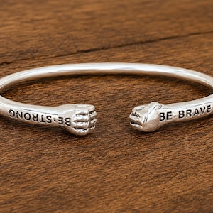 Op de afbeelding: Een zilverkleurige open armband met twee gebeeldhouwde vuisten aan de uiteinden. De tekst BE STRONG is aan de ene kant bij de vuist gegraveerd en de tekst BE BRAVE aan de andere kant bij de tweede vuist. De armband heeft een gladde afwerking.