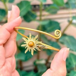 Könnte beinhalten: Ein goldfarbener Metall-Armreif mit einem zentralen sonnenförmigen Emblem, das in der Mitte einen runden, braunen, polierten Stein trägt. Das Armband hat ein Spiraldesign an einem Ende und ein texturiertes, gewickeltes Detail am Sonnenmotiv.