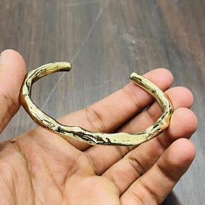 Op de afbeelding: Een goudkleurige metalen manchetarmband met een onregelmatig, organisch en getextureerd oppervlak. De armband heeft een open ontwerp met een gepolijste, reflecterende afwerking die het licht vangt en een ruwe, ongelijkmatige contour toont.