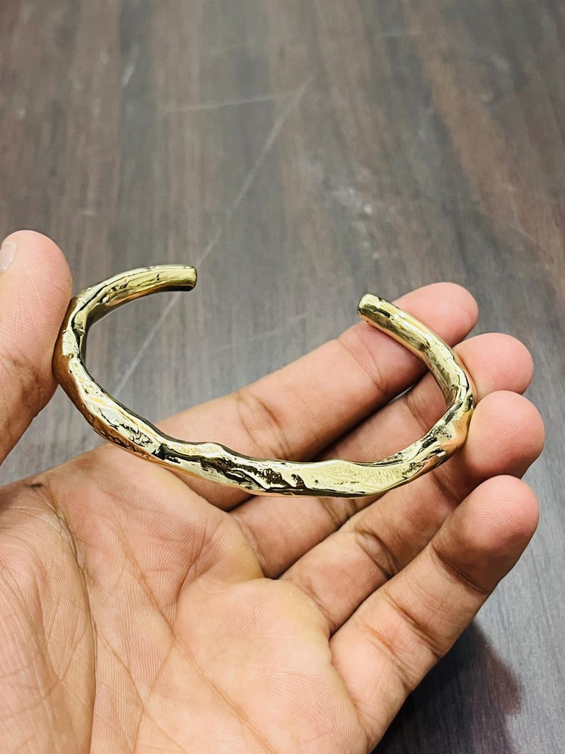 Op de afbeelding: Een goudkleurige metalen manchetarmband met een onregelmatig, organisch en getextureerd oppervlak. De armband heeft een open ontwerp met een gepolijste, reflecterende afwerking die het licht vangt en een ruwe, ongelijkmatige contour toont.