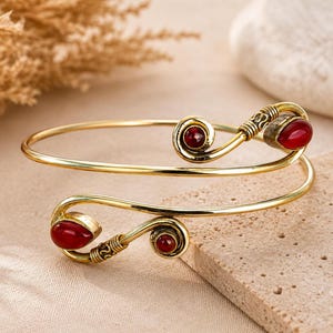 Peut inclure: Un bracelet de bras en métal doré présentant un design en spirale aux deux extrémités. Chaque extrémité est ornée d'une pierre rouge foncé en forme de goutte et d'une pierre ronde, avec des détails métalliques texturés près des sertis.