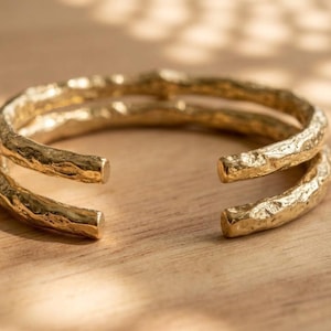 Op de afbeelding: Een goudkleurige manchetarmband met een dubbelbandig ontwerp en een getextureerd, onregelmatig oppervlak. De armband heeft een opening aan de voorzijde en toont een metallic afwerking die licht reflecteert op de ruwe, organisch ogende buitenkant.