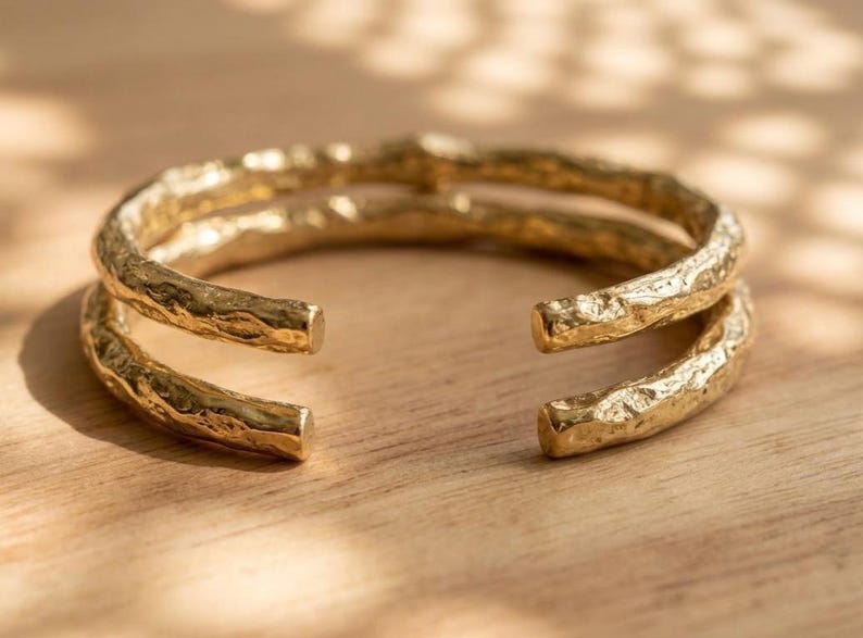 Op de afbeelding: Een goudkleurige manchetarmband met een dubbelbandig ontwerp en een getextureerd, onregelmatig oppervlak. De armband heeft een opening aan de voorzijde en toont een metallic afwerking die licht reflecteert op de ruwe, organisch ogende buitenkant.