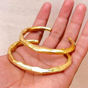 K&ouml;nnte beinhalten: Zwei goldfarbene Armreifen mit einer texturierten, unebenen Oberfl&auml;che. Jeder Armreif hat ein offenes Design mit einer polierten, reflektierenden Oberfl&auml;che, die das Licht einf&auml;ngt und eine Reihe organischer, unregelm&auml;&szlig;iger Vertiefungen erzeugt.