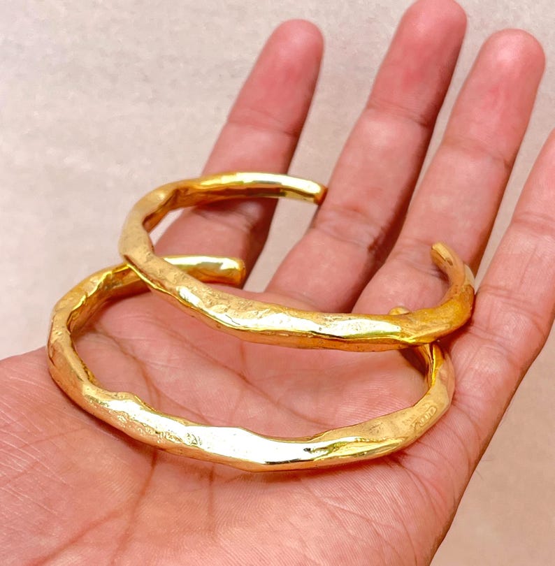 K&ouml;nnte beinhalten: Zwei goldfarbene Armreifen mit einer texturierten, unebenen Oberfl&auml;che. Jeder Armreif hat ein offenes Design mit einer polierten, reflektierenden Oberfl&auml;che, die das Licht einf&auml;ngt und eine Reihe organischer, unregelm&auml;&szlig;iger Vertiefungen erzeugt.