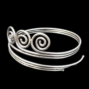 Könnte beinhalten: Ein Oberarmreif aus silberfarbenem Metall mit Spiraldesign. Das Schmuckstück besteht aus mehreren gewickelten Drahtbändern, die sich um den Arm legen, wobei drei markante kreisförmige Spiralmotive am oberen Rand des Reifs angebracht sind.