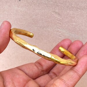 K&ouml;nnte beinhalten: Ein goldfarbenes Armband mit einer unregelm&auml;&szlig;igen, organischen und strukturierten Oberfl&auml;che. Das Metallband hat eine polierte Oberfl&auml;che, die das Licht auf seiner unebenen, wellenf&ouml;rmigen Form reflektiert und ein markantes, handgefertigtes Aussehen erzeugt.