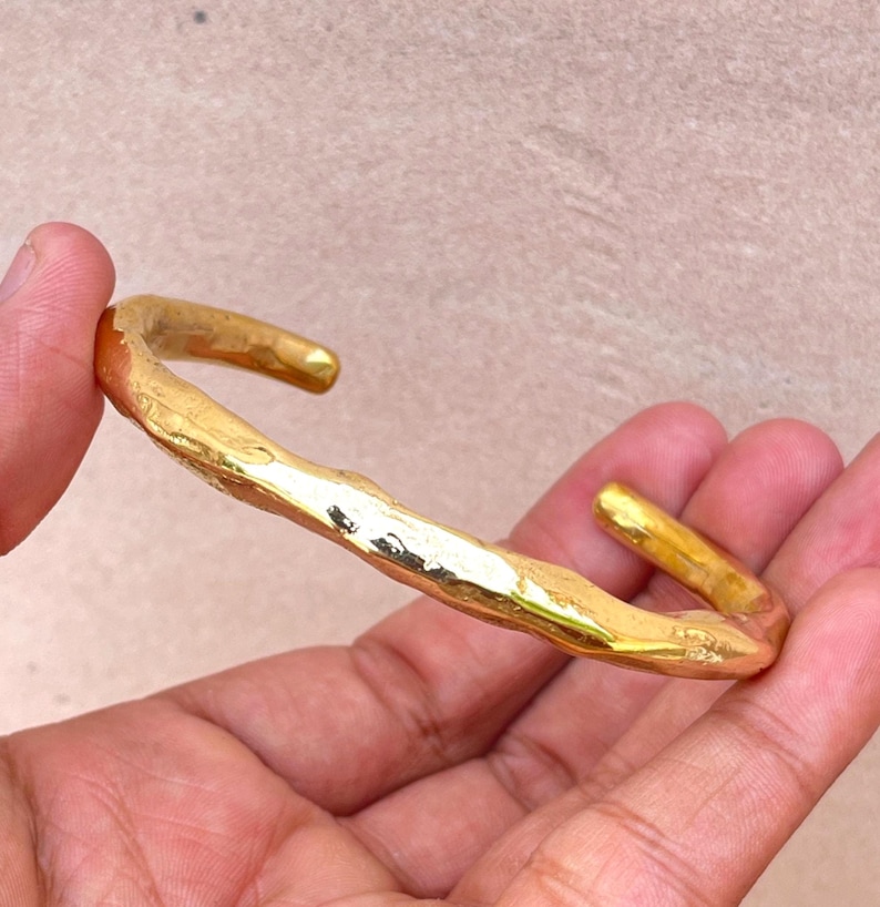 K&ouml;nnte beinhalten: Ein goldfarbenes Armband mit einer unregelm&auml;&szlig;igen, organischen und strukturierten Oberfl&auml;che. Das Metallband hat eine polierte Oberfl&auml;che, die das Licht auf seiner unebenen, wellenf&ouml;rmigen Form reflektiert und ein markantes, handgefertigtes Aussehen erzeugt.
