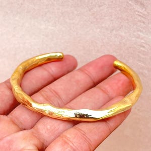 K&ouml;nnte beinhalten: Ein goldfarbenes Armband mit einer unregelm&auml;&szlig;igen, organischen und geh&auml;mmerten Textur. Das Armband hat ein offenes C-f&ouml;rmiges Design mit einer polierten, reflektierenden Oberfl&auml;che, die das Licht einf&auml;ngt und Highlights entlang der Kurve erzeugt.