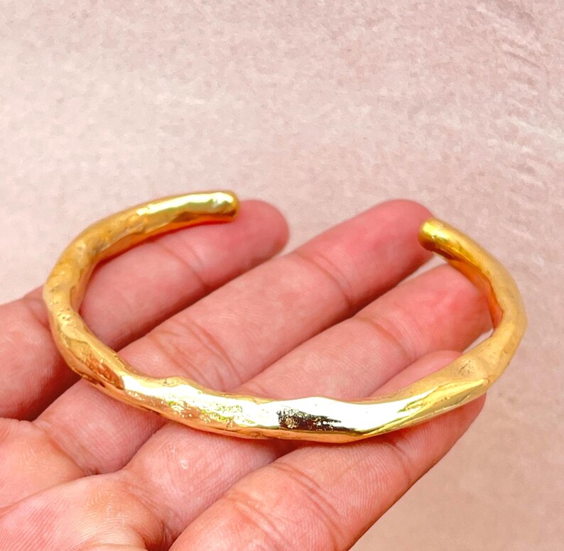 K&ouml;nnte beinhalten: Ein goldfarbenes Armband mit einer unregelm&auml;&szlig;igen, organischen und geh&auml;mmerten Textur. Das Armband hat ein offenes C-f&ouml;rmiges Design mit einer polierten, reflektierenden Oberfl&auml;che, die das Licht einf&auml;ngt und Highlights entlang der Kurve erzeugt.