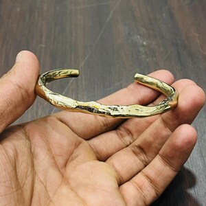 Op de afbeelding: Een goudkleurige metalen armband met een open ontwerp en een getextureerd, ongelijkmatig oppervlak dat het uiterlijk van organisch, ruw materiaal nabootst. De armband heeft een gepolijste afwerking die licht reflecteert op de onregelmatige vormen.