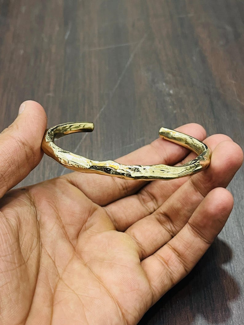 Op de afbeelding: Een goudkleurige metalen armband met een open ontwerp en een getextureerd, ongelijkmatig oppervlak dat het uiterlijk van organisch, ruw materiaal nabootst. De armband heeft een gepolijste afwerking die licht reflecteert op de onregelmatige vormen.