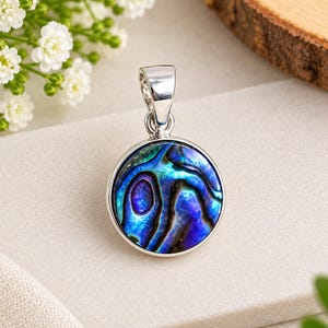 Könnte beinhalten: Ein runder Anhänger mit einer Einlage aus poliertem Abalone-Muschelmaterial, das irisierende Wirbel in tiefen Blau-, Violett- und Grüntönen aufweist. Die Muschel ist in einen glatten, glänzenden silberfarbenen Metallrahmen gefasst und hat eine Öse.