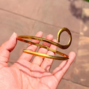 May include: A gold-toned metal arm cuff featuring a spiral design. The piece is constructed from a single continuous band that wraps around in a circular motion, ending in a curved, hook-like terminal at the top of the structure.