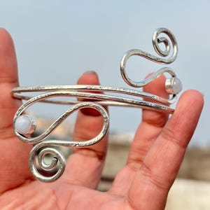 Puede incluir: Un brazalete de metal plateado con textura martillada y diseño abierto. Los extremos del brazalete terminan en espirales decorativas, cada una acentuada con una pequeña piedra preciosa blanca, redonda y translúcida en un engaste sencillo.