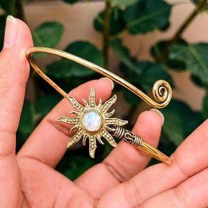 Peut inclure: Un bracelet manchette en métal doré orné d'un motif central en forme de soleil avec une pierre ronde blanche irisée en son centre. Le bracelet possède une tige ouverte lisse qui se termine par une spirale d'un côté et un motif solaire texturé.