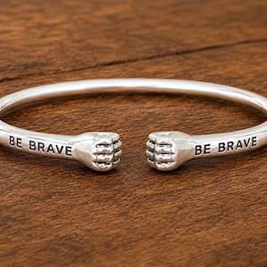 Op de afbeelding: Een zilverkleurige open armband met twee gebeeldhouwde vuistvormige uiteinden die naar elkaar toe wijzen. De woorden BE BRAVE zijn in zwarte letters gegraveerd aan beide zijden van de band, vlak voor de vuistdetails op dit metalen accessoire.