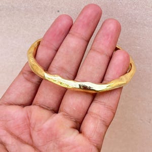 K&ouml;nnte beinhalten: Ein goldfarbener Armreif mit einer texturierten, unebenen Oberfl&auml;che. Das Metallband weist eine durchgehende, organische und unregelm&auml;&szlig;ige Form mit einer polierten, reflektierenden Oberfl&auml;che auf, die das Licht &uuml;ber den gesamten Umfang einf&auml;ngt.