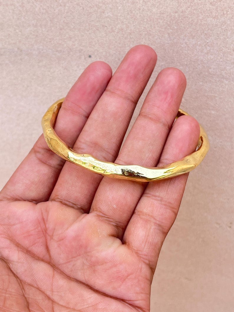 K&ouml;nnte beinhalten: Ein goldfarbener Armreif mit einer texturierten, unebenen Oberfl&auml;che. Das Metallband weist eine durchgehende, organische und unregelm&auml;&szlig;ige Form mit einer polierten, reflektierenden Oberfl&auml;che auf, die das Licht &uuml;ber den gesamten Umfang einf&auml;ngt.